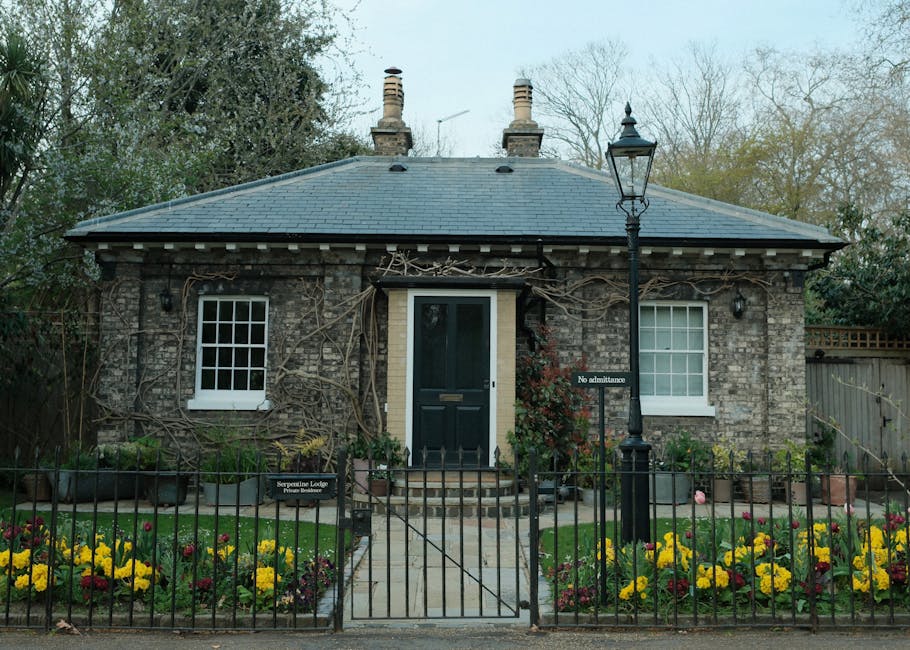 A small, single-story brick garden house with a dark tiled pitched roof, featuring two small white-framed windows and a black door at the center, accessible by a short set of brick steps. The exterior